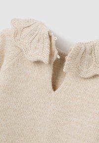 Cremig gestrickter Pullover mit einem gewellten Kragen, dekorativen Knöpfen und einem kleinen V-Ausschnitt. Weicher Stoff mit subtiler Eleganz.