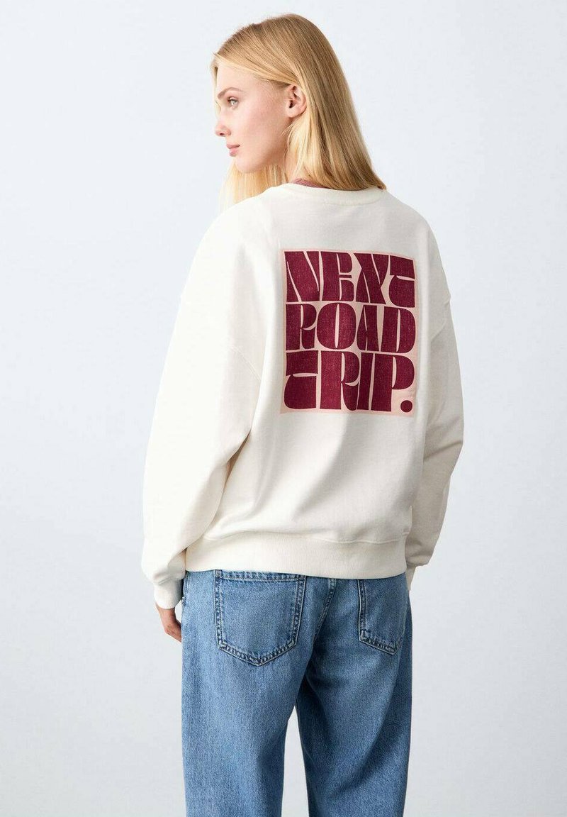 Sweatshirt de couleur crème avec un graphisme bordeaux "NEXT ROAD TRIP" au dos. Poignets et ourlet côtelés, avec une coupe décontractée.