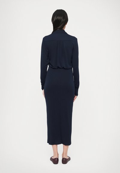 Rochie midi bleumarin cu mâneci lungi, guler și detalii drapate la spate. Țesătură moale, siluetă mulată și șlițuri laterale la tiv.