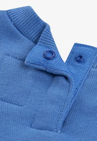 Blauer gestrickter Stoffkragen mit gerippter Textur, ausgestattet mit zwei Knopfverschlüssen und einem kleinen Taschen-Detail. Akzente in passenden Blautönen.