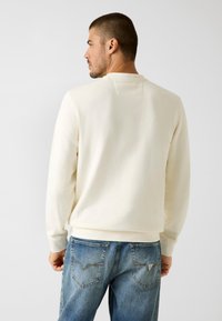 Crèmewitte sweatshirt met een ronde hals, geribbelde manchetten en zoom. Draagt lichtblauwe spijkerbroek met achterzakken en stikseldetails.