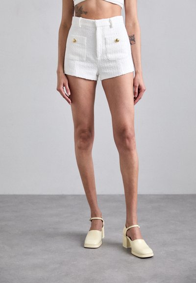 Shorts bianchi strutturati con tasche frontali e dettagli di bottoni dorati, abbinati a scarpe con tacco a blocco giallo pallido, su uno sfondo grigio.