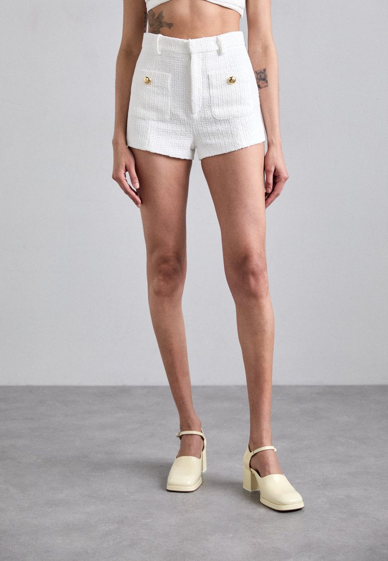 Shorts blancs texturés avec poches avant et accents de boutons dorés, associés à des chaussures à talons blocs jaunes pâles, sur fond gris.