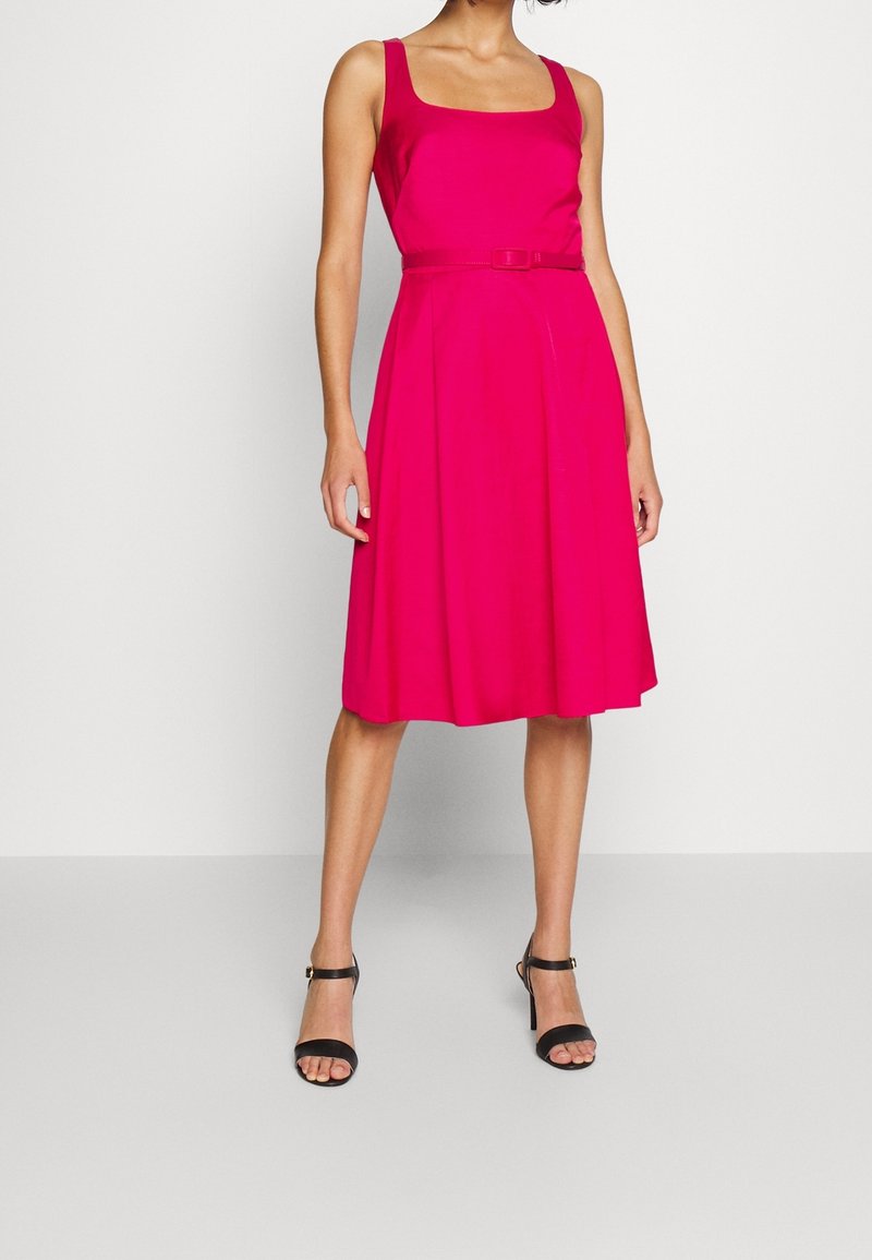 Fuchsiové bezramínkové šaty se čtvercovým výstřihem a páskem v pase; mají rozšířenou sukni a hladkou texturu. Černé sandály na podpatku dokončují vzhled.