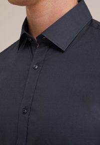 Chemise bleu marine à col classique. Présente une texture lisse, une fermeture à boutons avec un bouton noir, et des détails de couture visibles.