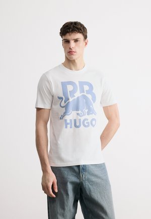 Jeune homme portant un T-shirt blanc avec un graphique de taureau bleu clair et le texte "RB HUGO", assorti à un jean en denim bleu clair, debout devant un fond blanc.