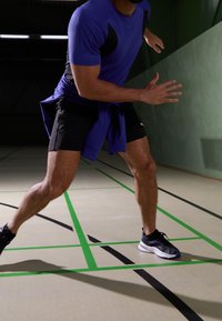 Conjunto atlético que incluye una camiseta ajustada morada y pantalones cortos negros. La persona está en movimiento en una cancha indoor texturizada, llevando zapatillas negras y azules.