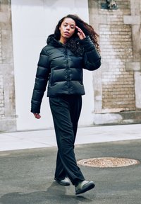 Schwarze Puffjacke mit Kapuze, ausgestattet mit einem gesteppten Design und voluminösen Ärmeln. Kombiniert mit dunklen Hosen und grünen Schuhen. Urbane Umgebung.