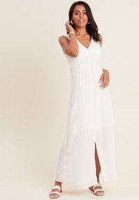 Robe midi blanche avec un décolleté en V, taille froncée et fente latérale. Fabriquée en tissu léger avec une texture subtile. Portée avec des sandales blanches.