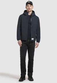 Marine vandtæt jakke med lynlås og elastiske manchetter, parret med mørke jeans og sorte sko. Accentueret af en kasket med et logo.
