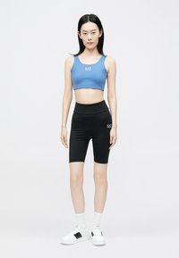 Kék sportos crop top logóval, magas derekú fekete kerékpáros rövidnadrággal párosítva. A modell fehér, fekete díszítéssel ellátott tornacipőt visel.
