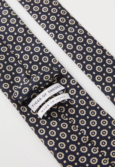 Mørkeblå slips laget av teksturert stoff med sirkulært og kvadratisk mønster i beige, med etiketter som sier "TIGER OF SWEDEN" og "MADE IN ITALY."