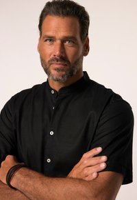 Musta kauluspaita lyhythihaisena, jossa on nappikiinnitys ja sileä pinta, päällä henkilö, jolla on kädet ristissä.