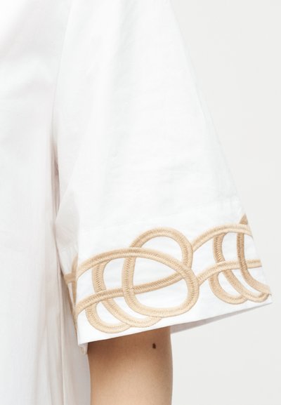 Chemise blanche à manches courtes avec des boucles beige brodées en chevauchement le long de l'ourlet visibles sur un bras avec un petit grain de beauté.