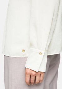 Weiße Bluse mit langen Ärmeln, verziert mit goldfarbenen Knöpfen an den Manschetten. Glatte Textur und lockere Passform, kombiniert mit hellvioletten Hosen.