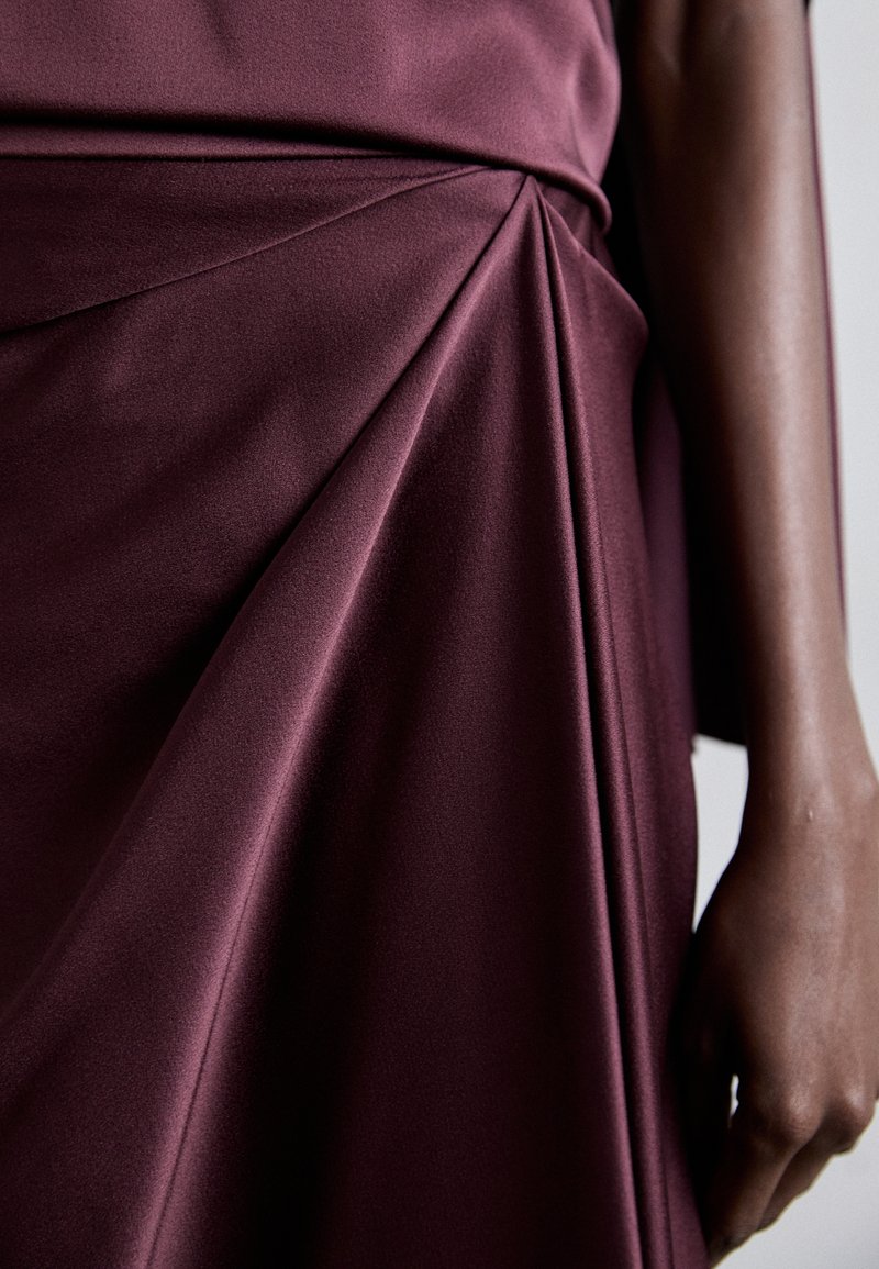 Burgunderfarvet kjole med blødt, glat stof, der har intrikate plisseringer og draperinger langs skørtet, hvilket fremhæver tekstur og bevægelse.