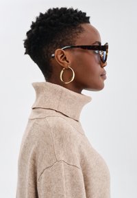 Beżowy sweter z golfem o prążkowanej fakturze, widok boczny, prezentujący czarne okulary przeciwsłoneczne i złote kolczyki na tle jednolitym.