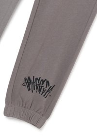 Pantalones de chándal grises hechos de una tela suave, con una cinturilla elástica y una impresión gráfica negra cerca del dobladillo. Textura suave, diseño casual.