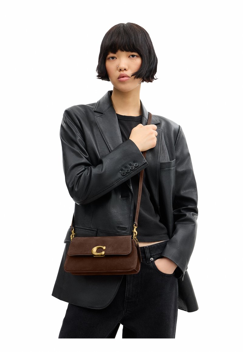 Jeune femme aux cheveux courts noirs portant une veste en cuir noire et un pantalon noir, tenant un sac à bandoulière en daim marron avec un fermoir doré.