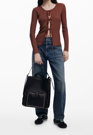 Osoba ubrana w brązowy prążkowany sweter z wiązaniami z przodu, niebieskie jeansy z wysokim stanem, czarne trampki, trzymająca czarną skórzaną torbę na ramię z przednią kieszenią.