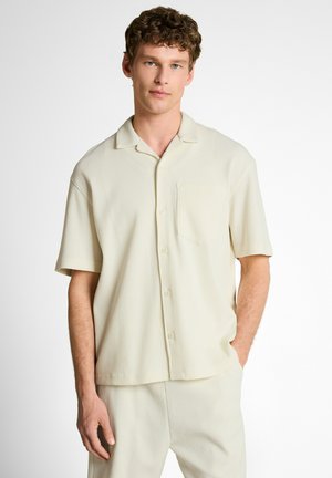 Młody mężczyzna ubrany w kremową koszulę na krótki rękaw zapinaną na guziki oraz pasujące spodnie, stojący z jedną ręką w kieszeni na białym tle.