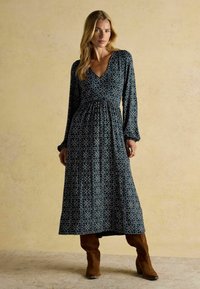Langärmliges, navy blaues Kleid mit einem sich wiederholenden Herzmuster und einem V-Ausschnitt. Hat eine geraffte Taille und einen ausgestellt Rock, kombiniert mit kniehohen braunen Stiefeln.