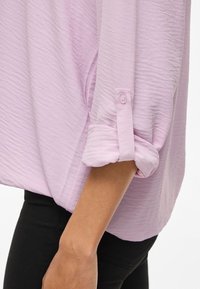 Blusa viola chiaro con tessuto testurizzato, caratterizzata da una manica arrotolata fissata da un bottone. Abbinata a pantaloni neri per contrasto.