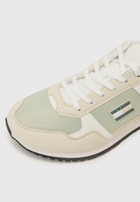 Scarpa sportiva beige e verde con materiali in suede e tessuto, lacci bianchi, suola piatta in gomma, piastrella con logo ben visibile sul lato.