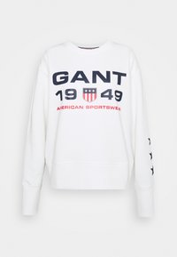 Sweatshirt blanc avec texte bleu marine et rouge, portant l'inscription "GANT 1949" et un design de bouclier. Manches plus longues avec des étoiles bleu marine sur un côté.