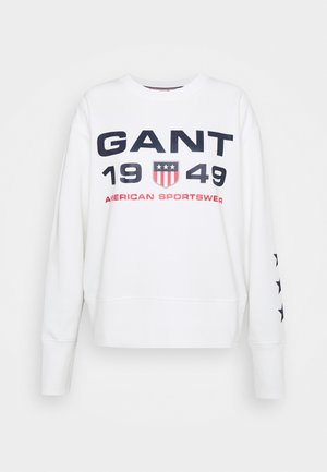 White sweatshirt with navy blue and red text, featuring "GANT 1949" and shield design. Longer sleeves with navy stars along one side.