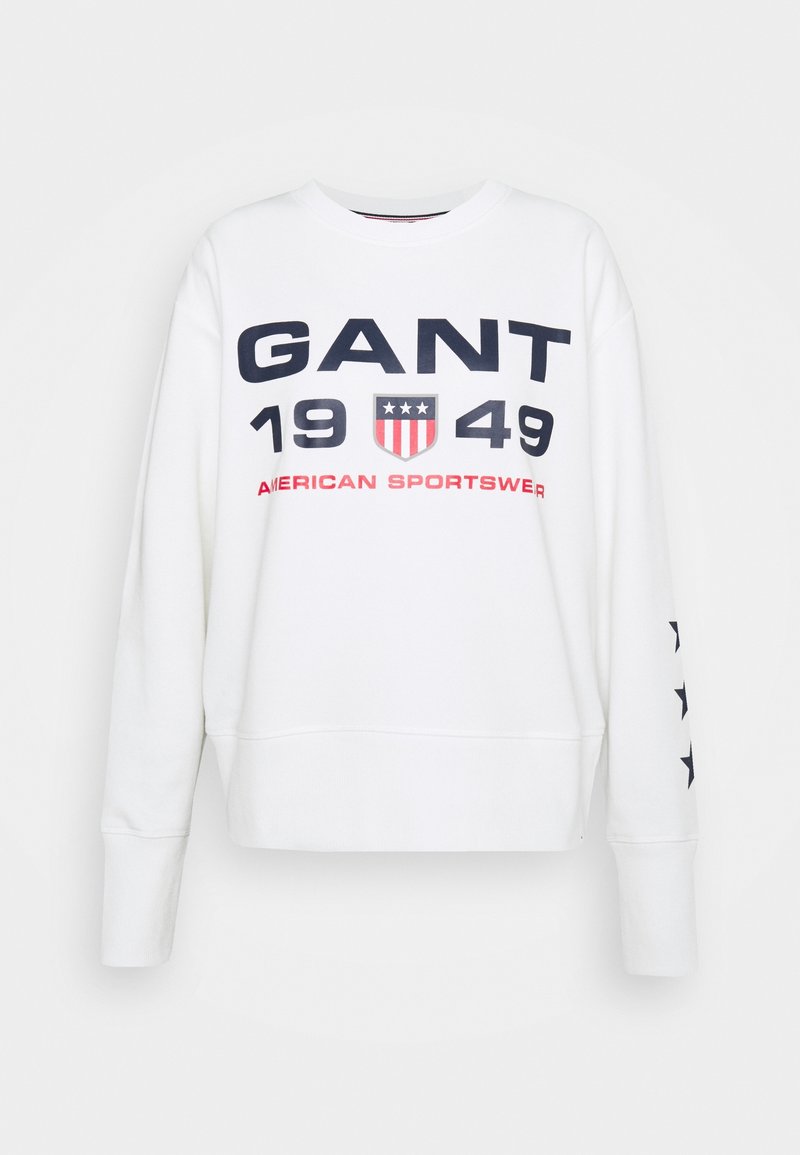 Sweatshirt blanc avec texte bleu marine et rouge, portant l'inscription "GANT 1949" et un design de bouclier. Manches plus longues avec des étoiles bleu marine sur un côté.