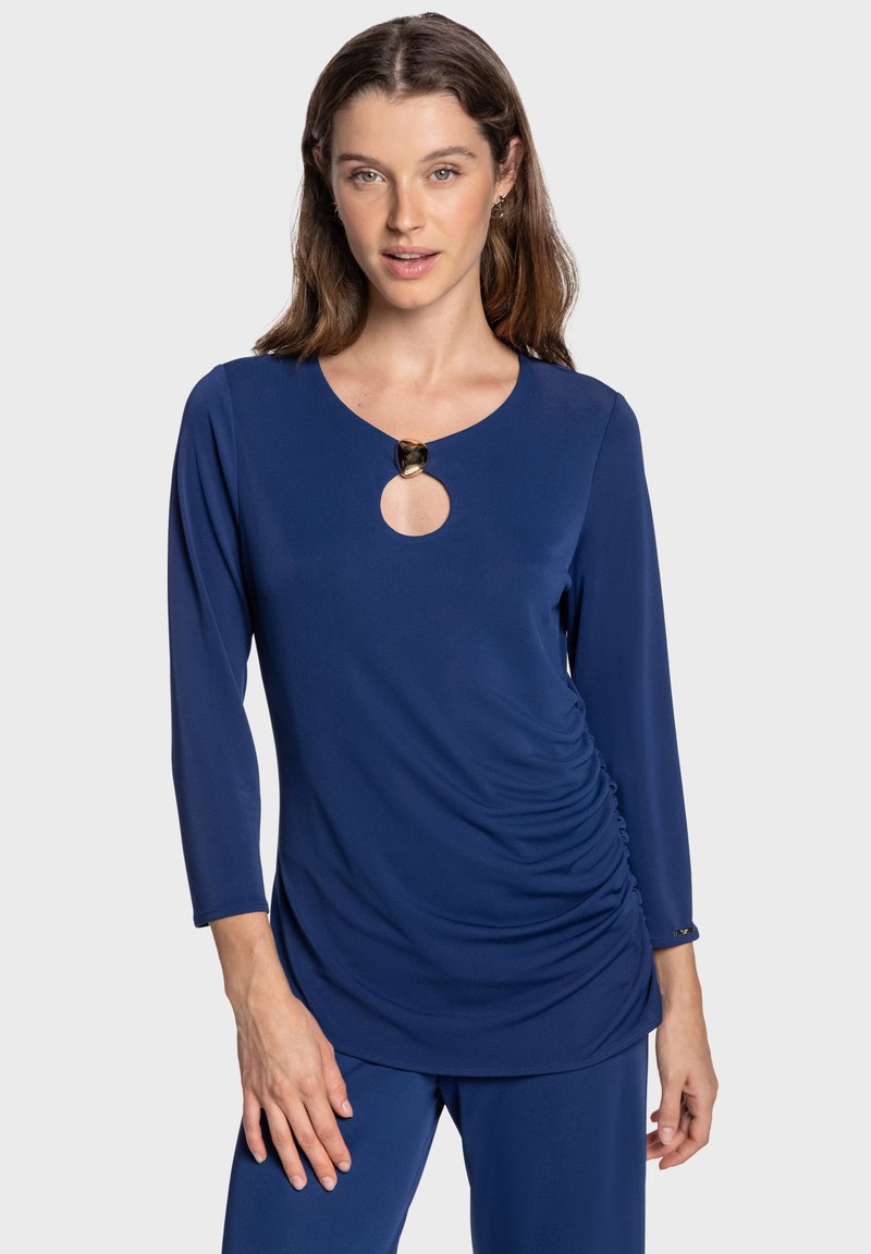 Haut à manches longues bleu marine avec un col rond, doté d'un détail en forme de goutte d'eau au niveau de la poitrine, de fronces sur les côtés et d'accents de garniture en métal doré.