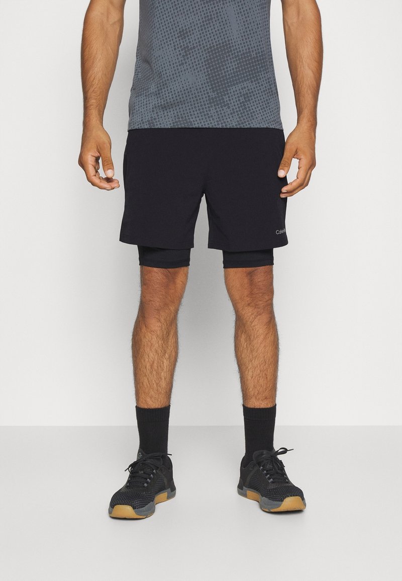 Shorts atletici neri con uno strato interno aderente, abbinati a una maglietta grigia a fantasia. Il modello indossa scarpe nere con suole in gomma, posando su uno sfondo bianco.