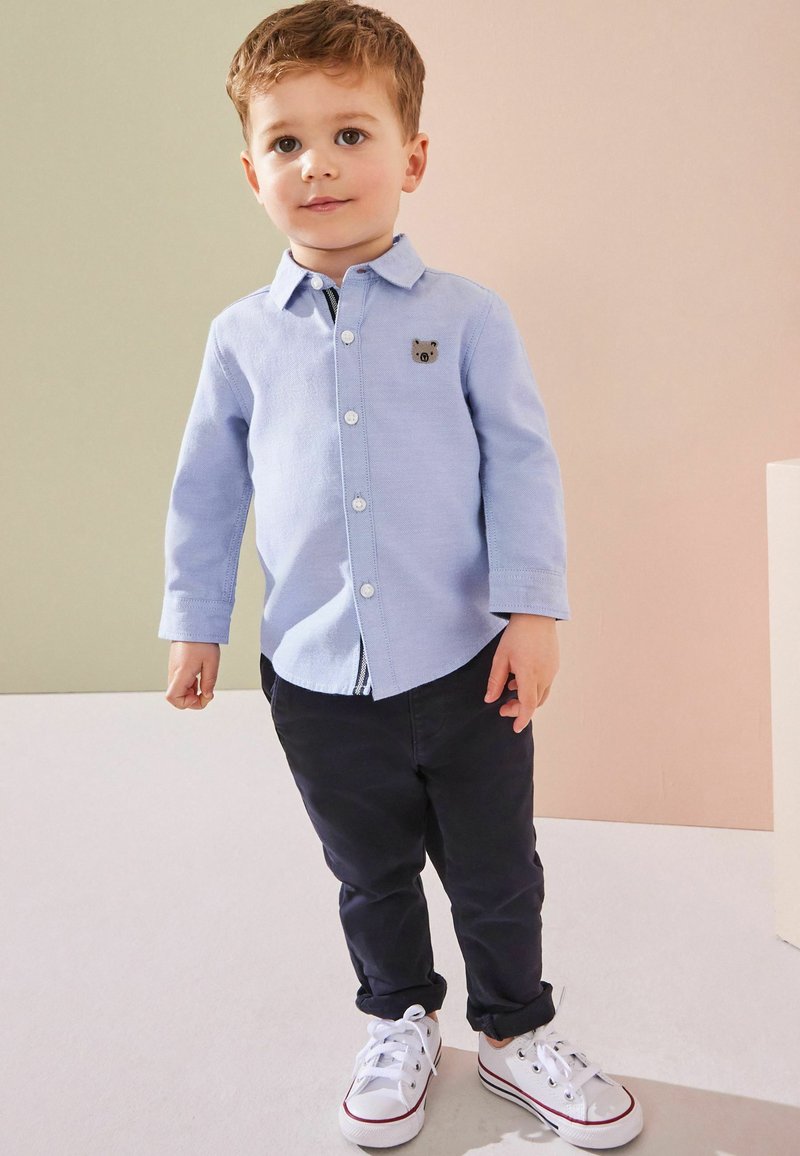 Camicia azzurro chiaro con bottoni e maniche lunghe, decorata da una toppa a forma di orso, abbinata a pantaloni blu scuro e scarpe da ginnastica bianche con dettagli rossi.
