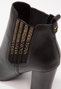 Bottine noire en cuir avec talon bloc, panneau latéral élastique orné de clous dorés verticaux, et languette arrière avec anneau métallique.