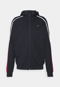 Marineblaues Zip-up Hoodie mit einer Kordelzugkapuze, das weiße, rote und blaue Streifen entlang der Ärmel und ein kleines Logo auf der Brust aufweist.