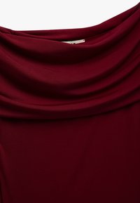 Tessuto borgogna drappeggiato con una texture morbida, caratterizzato da un collo a cascata. Il tessuto appare liscio con una leggera lucentezza.