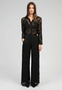 Chemisier en dentelle noire avec fermeture à boutons et manches longues, associé à un pantalon large en dentelle noire. Les deux vêtements présentent des motifs de dentelle intriqués.