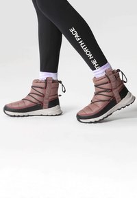 Botas de tobillo acolchadas rosas con suela negra, que presentan cordones negros y una correa con logotipo. Combinadas con leggings negros y calcetines morados.