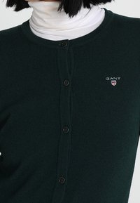 Dark green buttoned cardigan worn over a white turtleneck, featuring a small GANT logo on the left chest.