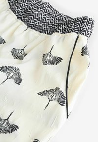 Leichtes Creme-Stoff mit einer zerknitterten Textur, auf dem schwarze Kranichmuster zu sehen sind. Der Bund hat ein schwarz-weißes geometrisches Design.