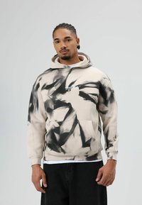 Beige capuchontrui met zwart tie-dye patroon, voorzien van een voorzak en een wit tekstlogo op de borst. Stof lijkt zacht.