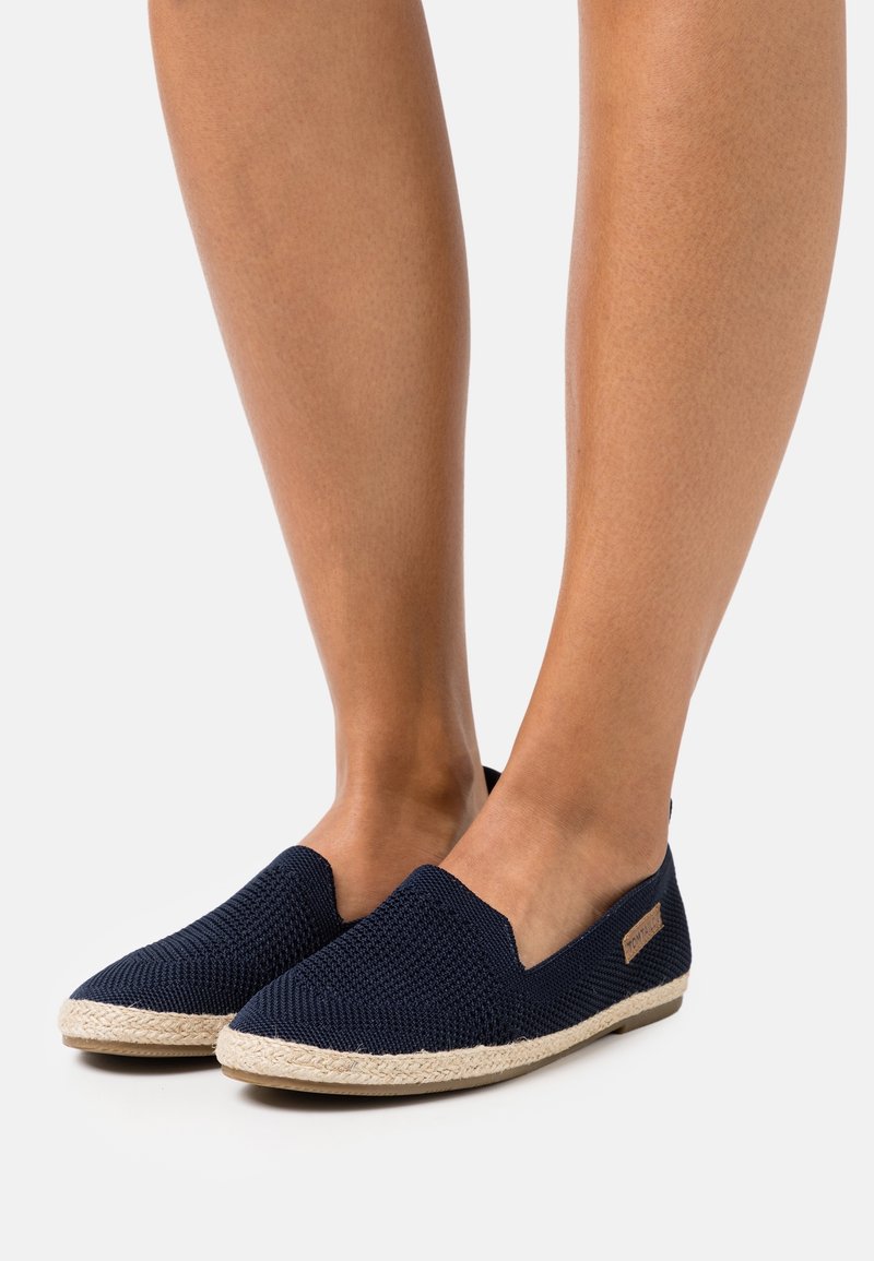 Jambes chaussées de chaussures à enfiler bleu marine en tissu texturé avec une bordure tissée beige le long de la semelle sur fond blanc.
