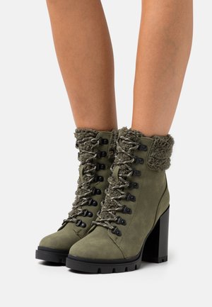 Olive green suede ankle boots with chunky heels, fur lining at the top, and lace-up front with metal eyelets worn on legs.