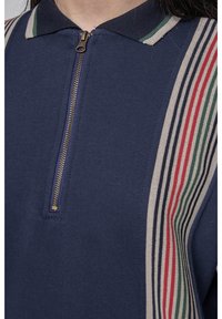 Sudadera marina con cuello con cremallera, que presenta rayas diagonales multicolores (rojo, verde, azul) a lo largo de los hombros. Textura de tela suave.