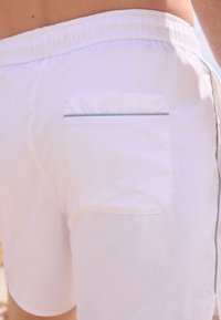 Hvide badeshorts lavet af letvægtsstof, med elastisk taljebånd, en baglomme med grå kant og en glat tekstur.