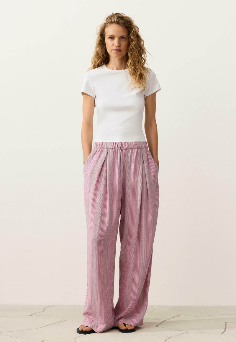 Vrouw staand, draagt een nauwsluitend wit kort mouwtopje, losse hoge taille roze gestreepte broek en zwarte sandalen, handen in de zakken.