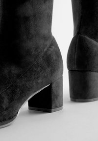 Juodos suede kulkšnies batai su kvadratiniu bloku aukštu, pasižymintys lygiu tekstūra ir paprastu dizainu. Aukštis suteikia stabilumo ir komforto.