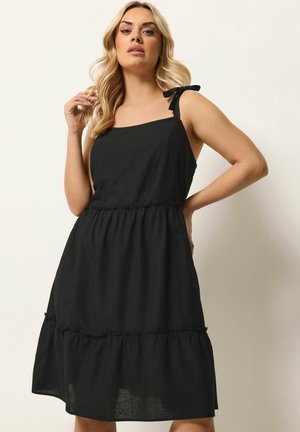 Femme blonde portant une robe noire sans manches avec des bretelles nouées aux épaules, debout devant un fond clair uni.