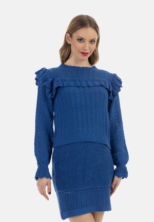 Donna che indossa un maglione blu lavorato a maglia a maniche lunghe con volant e una gonna blu lavorata a maglia abbinata, in piedi davanti a uno sfondo bianco.